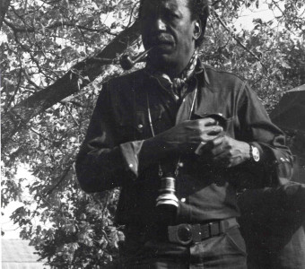 Director Gordon Parks is smoking a pipe as he reaches for his shirt pocket. A camera lens hangs from his neck.