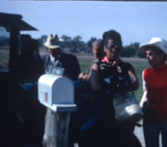 Actress Carol Lamond (Big Mabel), wearing round sunglasses, and other actors and crew prepare for a scene outside of Winger Home. The mailbox reads "Alex Mason."