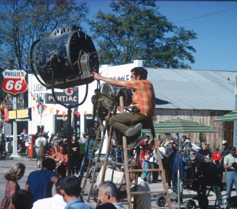 Actors and production crew filming in downtown Fort Scott, Kansas. One crew member is seated atop a tadder positioning a piece of lighting equipment.