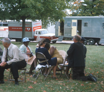 Actors, including Estelle Evans (Sarah Winger), obscured by actor Malcolm Attebury (Silas Newhall) and Don Dubbins (Harley Davis), far left, seated in front of production vehicles.