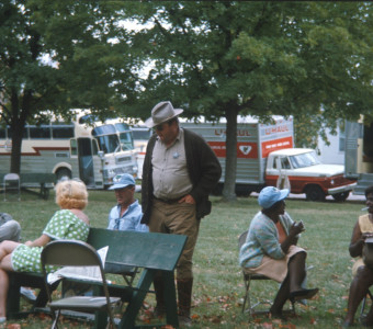 Actor Dana Elcar (Kirky) stands with seated cast members. All are photographed in front of production vehicles.