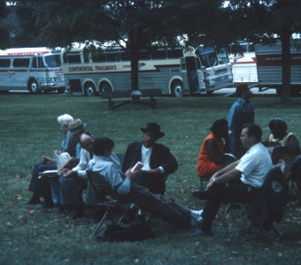 Acting cast, including Estelle Evans (Sarah Winger) on the far right, Stephen Perry (Jappy) standing in blue shirt and overalls, and Malcolm Atterbury (Silas Newhall) in black suit and hat, seated in front of production vehicles.