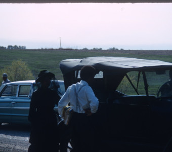 Actor Kyle Johnson (Newt Winger) and actress Hope Summers (Mrs. Kiner) stand left of black antique vehicle.