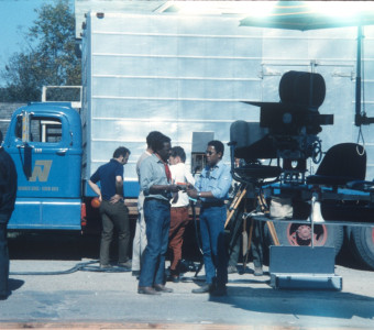 Director Gordon Parks, standing in the center of the photograph wearing a red ascot and blue jeans, speaks to his eldest son, Gordon Parks Junior, in front of production truck.