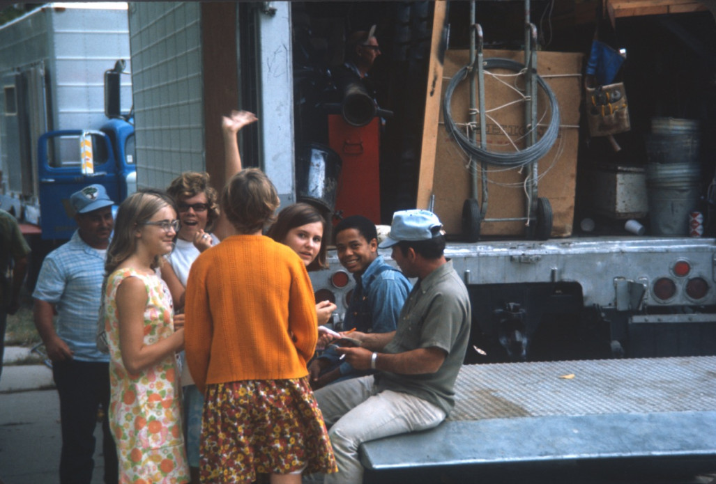 Crew, actors, and others, including Kyle Johnson (Newt Winger) wearing blue denim overalls, mingling in front of production equipment. Johnson appears to be signing autographs for the young ladies in the picture.