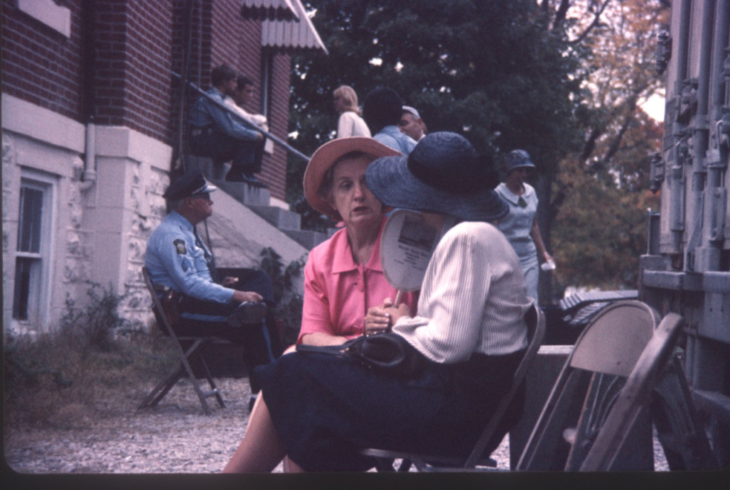 Actors and crew mingle outside of courthouse building. Two actresses talk while security officer sits next to courthouse stairs.