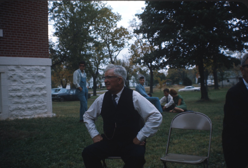 Actor wearing black suit vest seated outside of courthouse building. Additional actors seated in background.