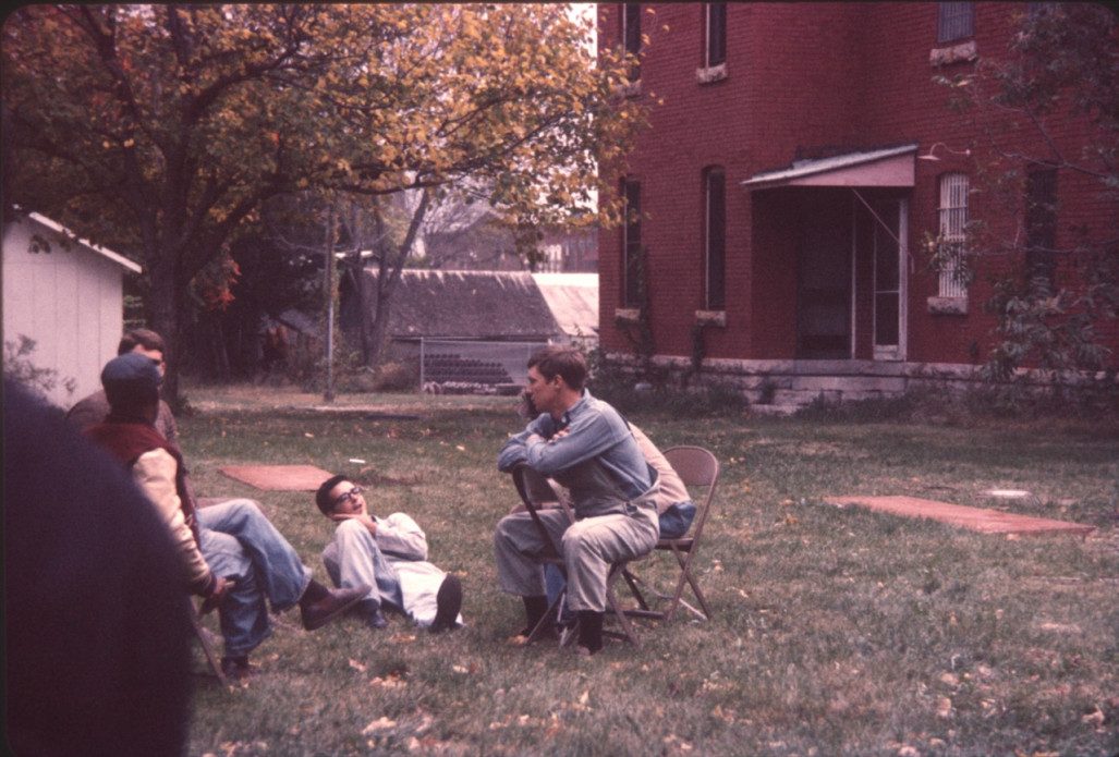 Five actors talking in the grass beside the courthouse building from trial scene.