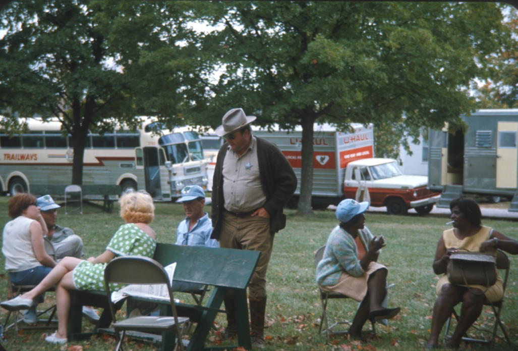 Actor Dana Elcar (Kirky) stands with seated cast members. All are photographed in front of production vehicles.