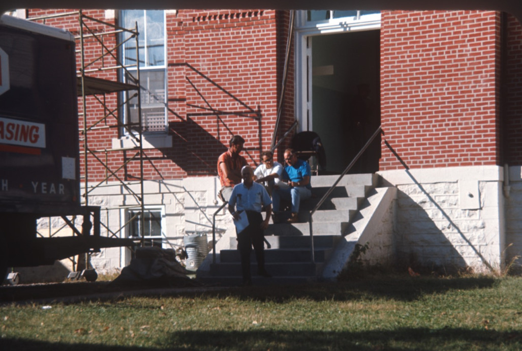 Crew members and production equipment outside of courthouse building from trial scene.