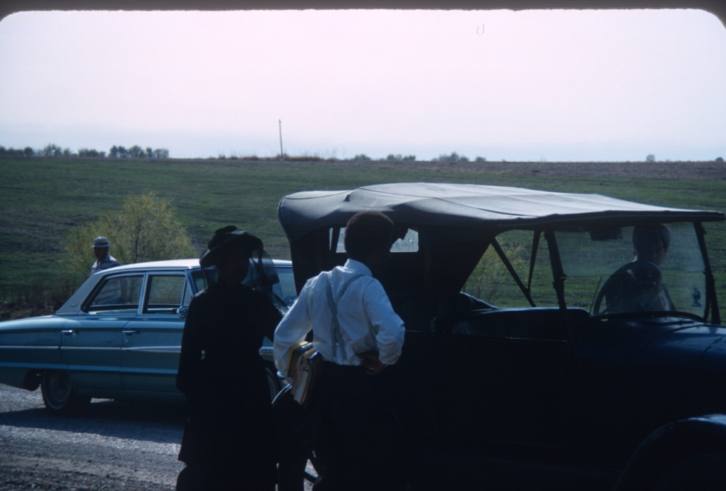 Actor Kyle Johnson (Newt Winger) and actress Hope Summers (Mrs. Kiner) stand left of black antique vehicle.