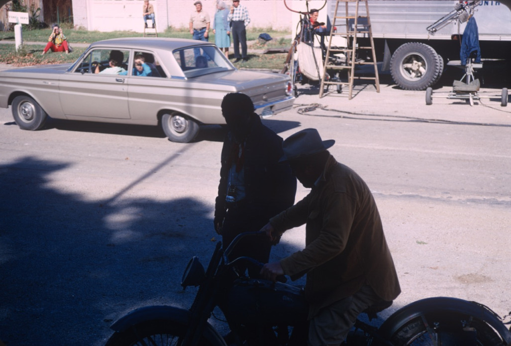 Director Gordon Parks stands alongside actor Dana Elcar (Kirky) who is seated on a motorcycle in front of production crew and equipment.