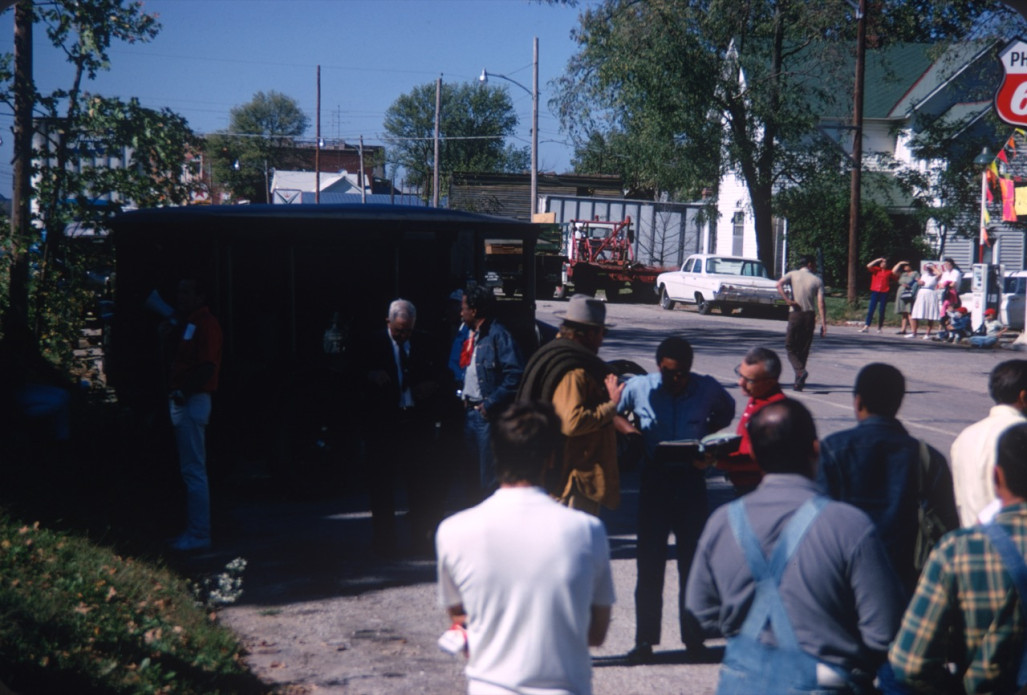 Actors Dana Elcar (Kirky) and Kyle Johnson (Newt Winger) consult book with a crew member with production crew and cast around them. Director Gordon Parks standing behind them in red ascot in front of hearse. Photograph location in downtown Fort Scott, Kansas.